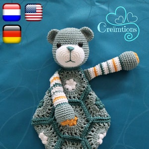 May include: Crocheted bear security blanket in shades of gray, white, and orange. The bear has a white face, a teal nose, and a flower on its chest. The blanket is hexagonal with a lacy design. Text on the image reads "Kleine-Knuffel-Beer".