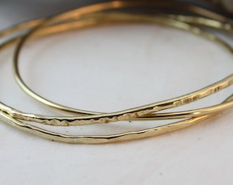 May include: Three gold bangles stacked on top of each other. The bangles are smooth and have a hammered texture.