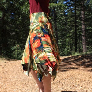 May include: A woman wearing a colorful, abstract print skirt with a high waist and a flowing, asymmetrical hem. She is standing in a forest setting with trees in the background.