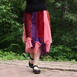 Puede incluir: Una mujer con una falda multicolor, un top negro y zapatos negros. La falda está hecha de diferentes telas y tiene un dobladillo con volantes. La mujer está de pie en un camino de ladrillos.