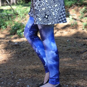 May include: Blue galaxy print leggings with white clouds and stars, worn with a black and white patterned top and black flats. The photo is taken outdoors in a forest setting. The leggings are a striking fashion item.