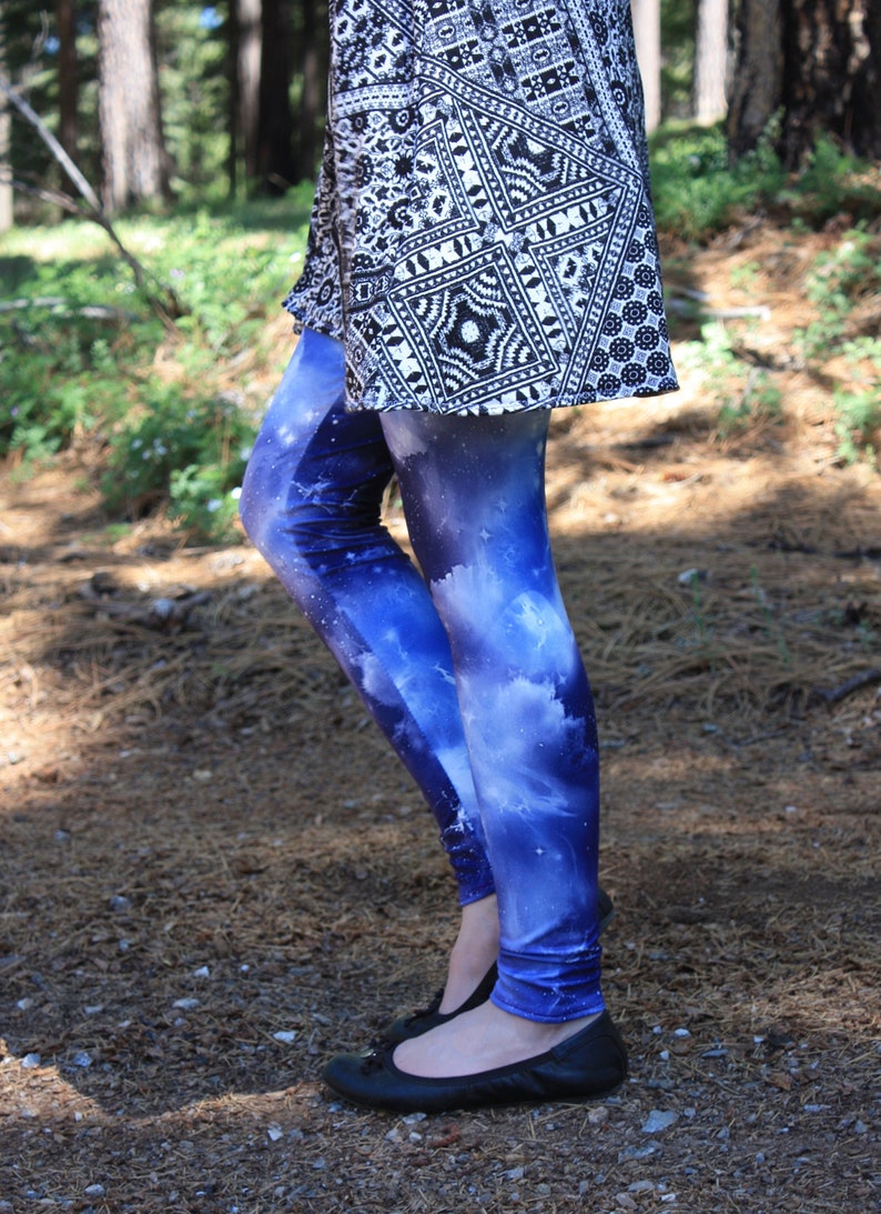 May include: Blue galaxy print leggings with white clouds and stars, worn with a black and white patterned top and black flats. The photo is taken outdoors in a forest setting. The leggings are a striking fashion item.