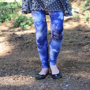 May include: Blue galaxy-print leggings paired with a black and white patterned skirt. The leggings display a vibrant cosmic design in blue, purple, and white. Black ballet flats are worn. The image is taken outdoors.
