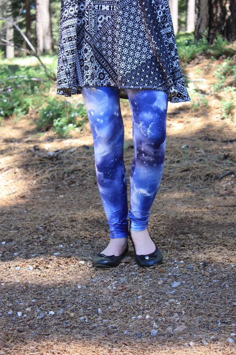 May include: Blue galaxy-print leggings paired with a black and white patterned skirt. The leggings display a vibrant cosmic design in blue, purple, and white. Black ballet flats are worn. The image is taken outdoors.