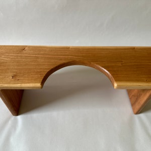 May include: A wooden footstool with a natural finish. The stool has a rectangular top with a curved cutout in the center and angled legs. The wood grain is visible, and the stool is approximately 24 inches long.