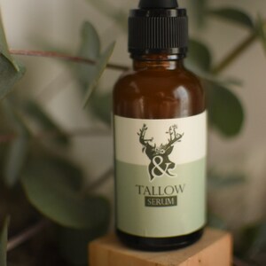 May include: A brown glass bottle with a black dropper and a white and green label. The label features a deer illustration and the words "& Tallow Serum". The bottle is displayed on a small wooden block.
