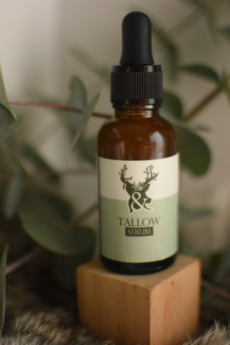 May include: A brown glass bottle with a black dropper and a white and green label. The label features a deer illustration and the words "& Tallow Serum". The bottle is displayed on a small wooden block.
