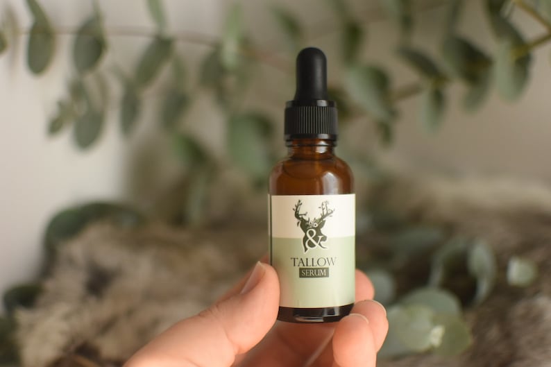 May include: A small amber glass bottle with a black dropper top, labeled "Tallow Serum." The label features a deer graphic and a light green and white color scheme. The bottle is held in a hand, with a blurred background of greenery.