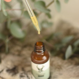 May include: A brown glass bottle with a dropper dispensing a yellow liquid. The bottle has a white label with a green band and a deer emblem. The background includes greenery and a soft, textured surface.