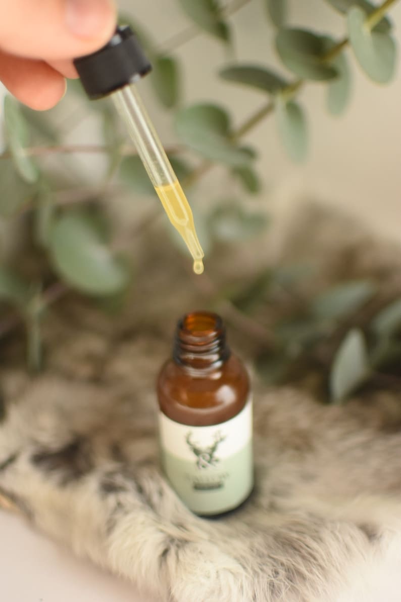 May include: A brown glass bottle with a dropper dispensing a yellow liquid. The bottle has a white label with a green band and a deer emblem. The background includes greenery and a soft, textured surface.