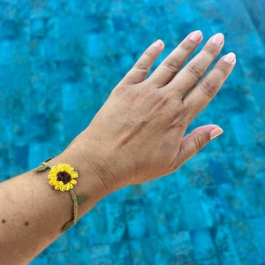 May include: A crochet bracelet featuring a yellow and brown sunflower design. The sunflower is the focal point, with green leaves and a green vine-like band. The bracelet is worn on a wrist against a blue background.