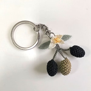 May include: A silver key chain with a crocheted olive branch charm. The charm features three olives, one black, one green, and one brown, and a white flower with a yellow center.