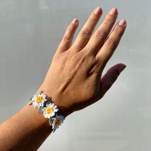 May include: A daisy bracelet featuring white petals, yellow centers, and green leaves. The floral design is crafted with intricate detail, showcasing a delicate and feminine aesthetic. The bracelet is a charming accessory.