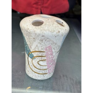 Puede incluir: Un portacepillos de cerámica moteada con tres agujeros. El soporte presenta diseños abstractos dorados, azules y rosas. El soporte es de color beige claro.