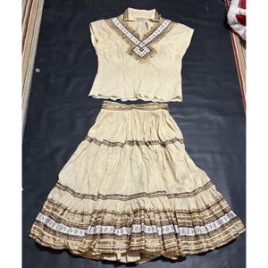 May include: A two-piece outfit consisting of a short-sleeved cream blouse and a matching tiered skirt. Both pieces feature intricate brown and white embroidered patterns along the neckline, bodice, and horizontal bands on the skirt fabric.