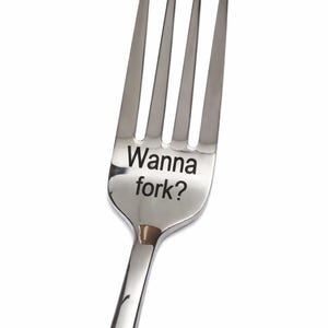 May include: A stainless steel fork with four tines. The words "Wanna fork?" are etched into the fork's head. The fork is set against a white background.
