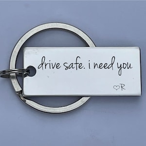 May include: A rectangular silver keychain with a large ring. The white tag has the words "drive safe. I need you" in black script. A small heart and the letter "R" are at the bottom right.