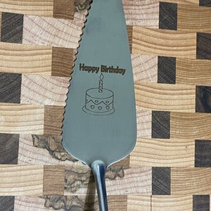 May include: A silver cake server with a serrated edge and a handle. The server is engraved with the words "Happy Birthday" and a birthday cake with a candle.