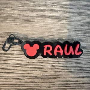 May include: A black and red keychain with the name "RAUL" in red letters. The design includes a red Mickey Mouse head silhouette on the left side. The keychain has a black clasp.