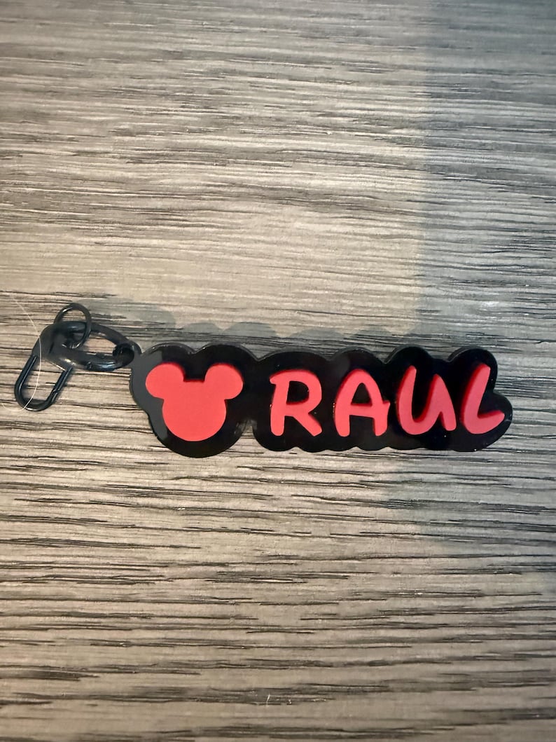 May include: A black and red keychain with the name "RAUL" in red letters. The design includes a red Mickey Mouse head silhouette on the left side. The keychain has a black clasp.