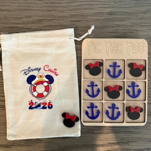 Puede incluir: Un juego de tic-tac-toe de madera con cabezas de Minnie Mouse negras con lazos rojos y anclas azules. Una bolsa de lona con el logotipo de Disney Cruise y el año 2020 está al lado del juego. Una cabeza de Minnie Mouse está al lado de la bolsa.