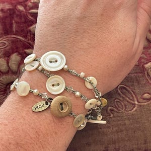 May include: A bracelet made of multiple strands of beads and buttons. The bracelet features a variety of white and brown buttons, including some with a pearl-like finish. The bracelet also includes a small silver tag with the text "FDM" on it.
