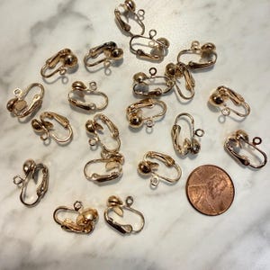 May include: A collection of gold-toned metal clip-on earring findings, each featuring a small loop for attachment and a hinged mechanism. A single copper-colored United States penny is included in the arrangement for size comparison.