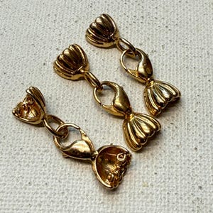 May include: Three gold-colored jewelry clasps with a decorative, shell-like design. Each clasp features a lobster claw closure and is connected to a decorative end piece. The clasps are arranged on a textured, off-white surface.