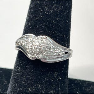 May include: A silver ring with a cluster of small, clear stones set in a decorative design. The ring is displayed on a black velvet surface, showcasing its intricate details and sparkling appearance. The ring's band has a split design.