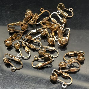 May include: A pile of gold-toned earring clips. The clips are made of metal and have a small ball on one end. They are designed to be used with earrings that do not have a post.
