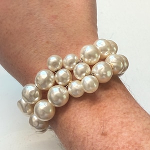 May include: A cream-colored pearl bracelet. The bracelet features multiple strands of large, round, faux pearls. The pearls are strung together with gold-colored metal links. The bracelet is worn on a wrist.