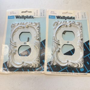 May include: Two vintage Sears Wallplate outlet covers. The covers are white with ornate, raised scrollwork and gold accents. Each is packaged on a blue and white card. The text "Sears Wallplate" is printed on the card.