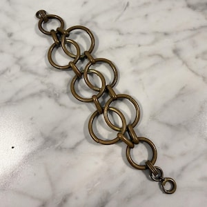 May include: A vintage-style bracelet featuring a series of interconnected bronze-colored rings. The bracelet has a clasp closure and is displayed on a marble surface. The design is simple yet elegant, suitable for various occasions.