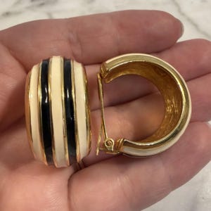 May include: A pair of gold-tone hoop earrings featuring alternating vertical stripes of cream and black enamel. The earrings have a thick, rounded profile and include a hinged clip-on mechanism on the back for secure attachment to the earlobe.