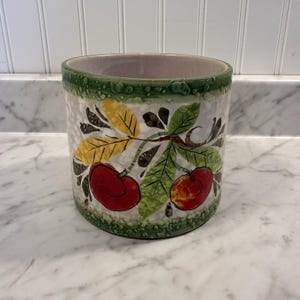May include: A cylindrical ceramic container with a green rim and a white background. The container is decorated with a branch of red cherries and green and yellow leaves. The rim has a textured, dotted pattern.