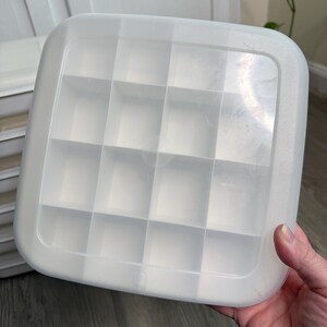 May include: A clear, square plastic container with a grid of 16 compartments. The container has rounded corners and is held by a hand. The lid is translucent and appears to be made of a durable plastic material. The container is designed for organizing small items.