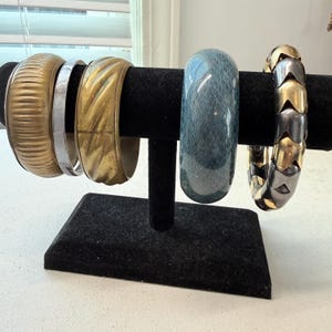 May include: A collection of bracelets displayed on a black velvet stand. The bracelets include gold-toned, silver, and a blue patterned bangle. One bracelet has a textured surface, while another has a snake-like pattern. The bracelets are of varying widths and styles.