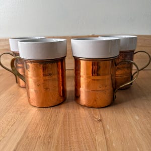 May include: Four copper mugs with white ceramic rims and brass handles are arranged on a wooden surface. The mugs have a cylindrical shape and a polished finish, reflecting the light. The handles are curved and add a touch of elegance.
