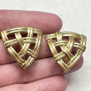 May include: A pair of gold-toned clip-on earrings. Each earring is a triangle shape with a woven basket design. The earrings are made of a shiny, metallic material.