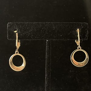 May include: A pair of gold-toned hoop earrings. Each earring features a circular design suspended from a lever-back closure. The earrings are displayed against a black background, highlighting their metallic sheen and simple elegance.