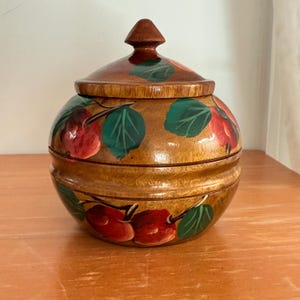 May include: A wooden jar with a lid, painted with red cherries and green leaves. The jar is brown with a dark brown lid and finial. The jar is round with a two-part design.