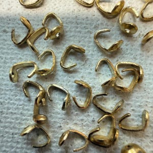 May include: Close-up of several small, gold-colored jewelry findings. The findings are a variety of shapes, including curved clasps and connectors, scattered across a white surface. These components are likely used in jewelry making and crafting.