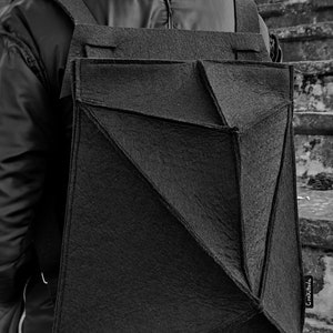 May include: Black geometric backpack with a unique angular design. The backpack is made of a textured material and features adjustable straps. The bag is worn over the shoulders. The background is a set of concrete steps.