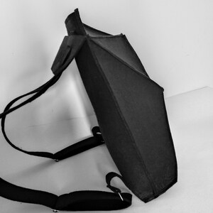 May include: Black backpack with adjustable shoulder straps. The backpack has a geometric design with a rectangular body and a triangular top. The straps and the body of the backpack are made of a dark material.