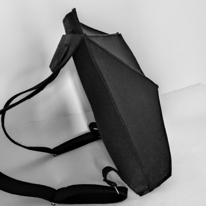 May include: Black backpack with adjustable shoulder straps. The backpack has a geometric design with a rectangular body and a triangular top. The straps and the body of the backpack are made of a dark material.