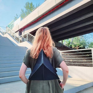 Peut inclure: Une personne portant une robe verte avec un sac à dos triangulaire noir. La personne est debout sur un escalier en béton sous un pont.