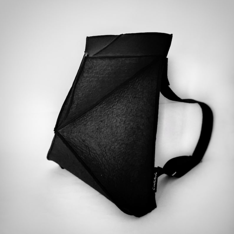 May include: Black geometric shoulder bag with a textured surface and a long, adjustable strap. The bag has a unique, angular design and a small tag with text. The bag is set against a white background.