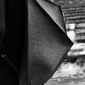 May include: Black, geometric-shaped shoulder bag with a textured surface. The bag is worn over a black sweater and dark jeans. The background is a blurred outdoor scene.