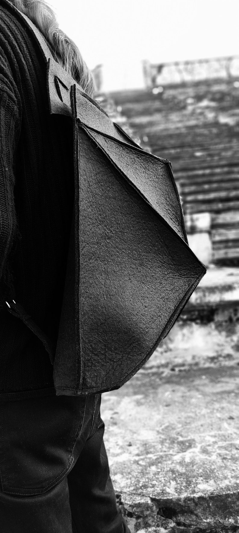 May include: Black, geometric-shaped shoulder bag with a textured surface. The bag is worn over a black sweater and dark jeans. The background is a blurred outdoor scene.