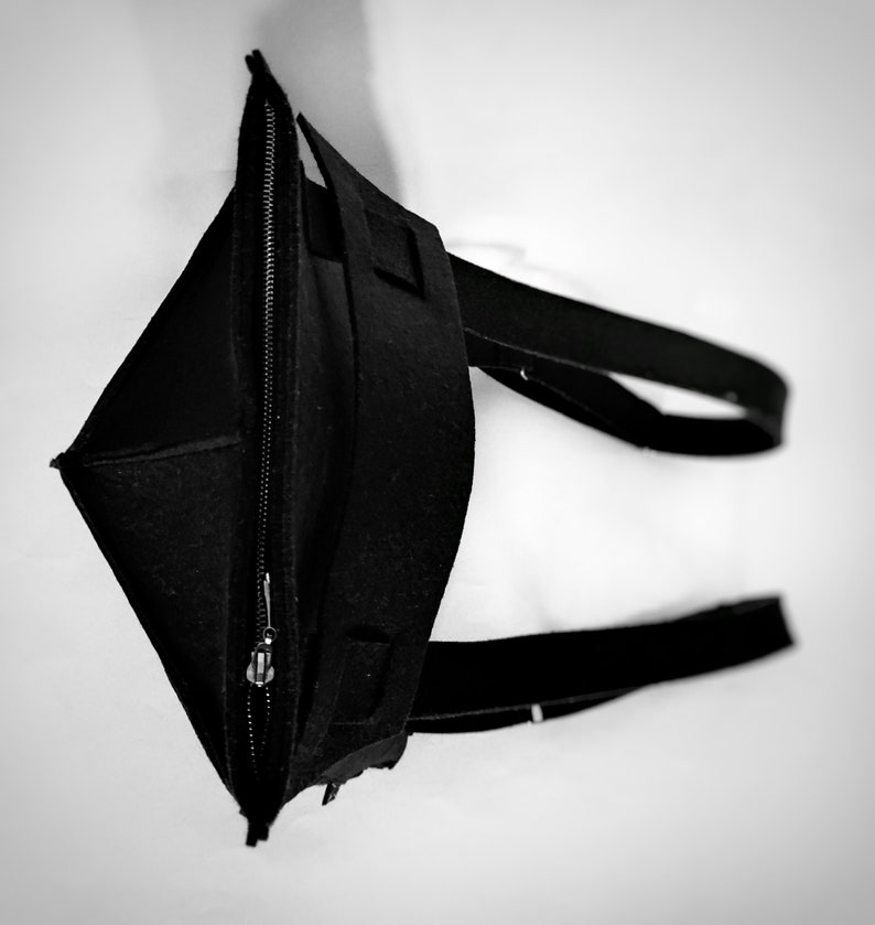 May include: A black tote bag made of felt, featuring a zippered closure and two long straps. The bag is open, revealing the interior. The design is simple and modern, with a focus on functionality and a minimalist aesthetic.